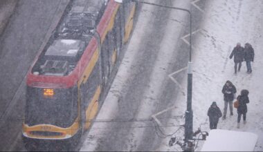 Tramwaj porusza się w trudnych warunkach pogodowych podczas opad&oacute;w śniegu, a na poboczu stoją przechodnie.