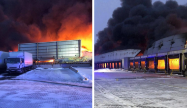 Pożar hali magazynowej z gęstym dymem i płomieniami, widoczna ciężar&oacute;wka i doki załadunkowe.