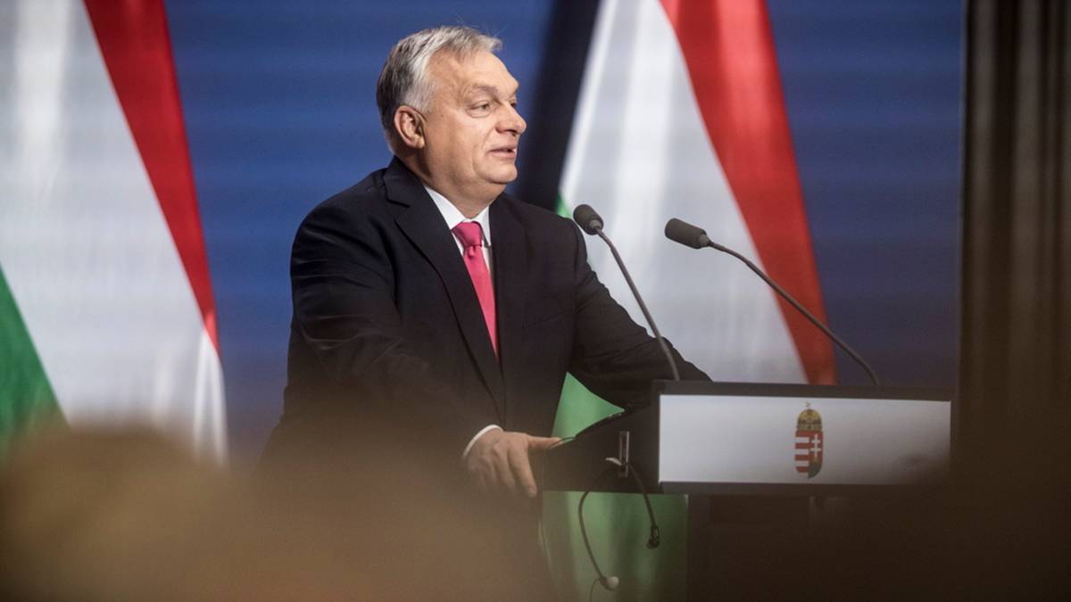 Mężczyzna w garniturze przemawia za m&oacute;wnicą z herbem Węgier, w tle flagi Węgier.