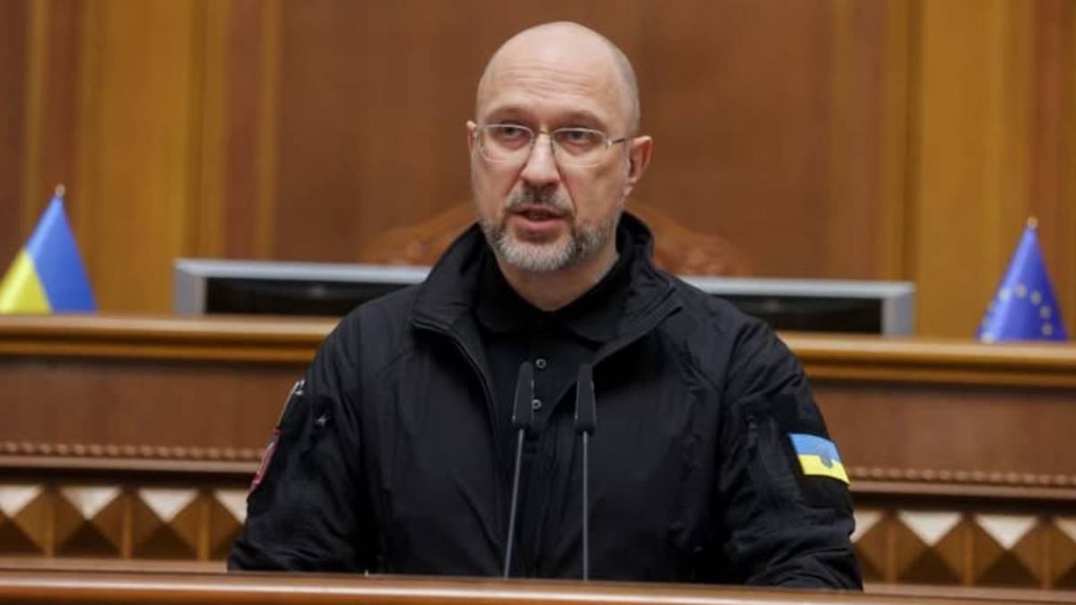 Mężczyzna w czarnej kurtce z naszywkami z flagą Ukrainy i okularami siedzi za m&oacute;wnicą z mikrofonami.