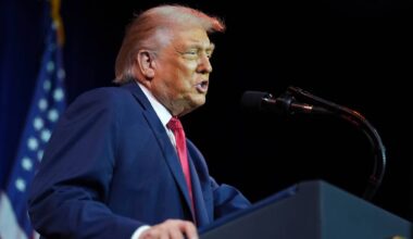 Donald Trump przemawia za m&oacute;wnicą, obok niego znajduje się mikrofon, w tle powiewa amerykańska flaga.