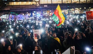 Tłum ludzi trzyma w g&oacute;rze światła telefon&oacute;w podczas nocnej demonstracji, na kt&oacute;rej widoczna jest tęczowa flaga.