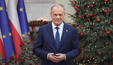 Premier Donald Tusk przemawia na tle flag Polski i Unii Europejskiej oraz świątecznej choinki.