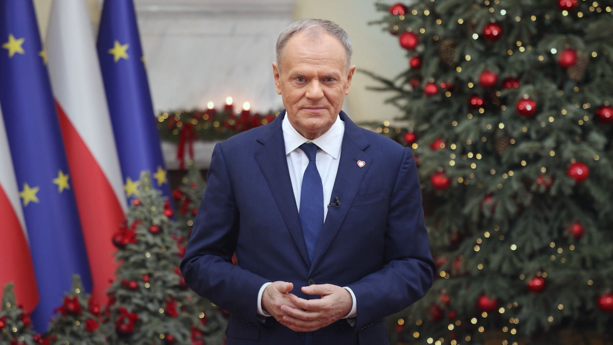 Premier Donald Tusk przemawia na tle flag Polski i Unii Europejskiej oraz świątecznej choinki.