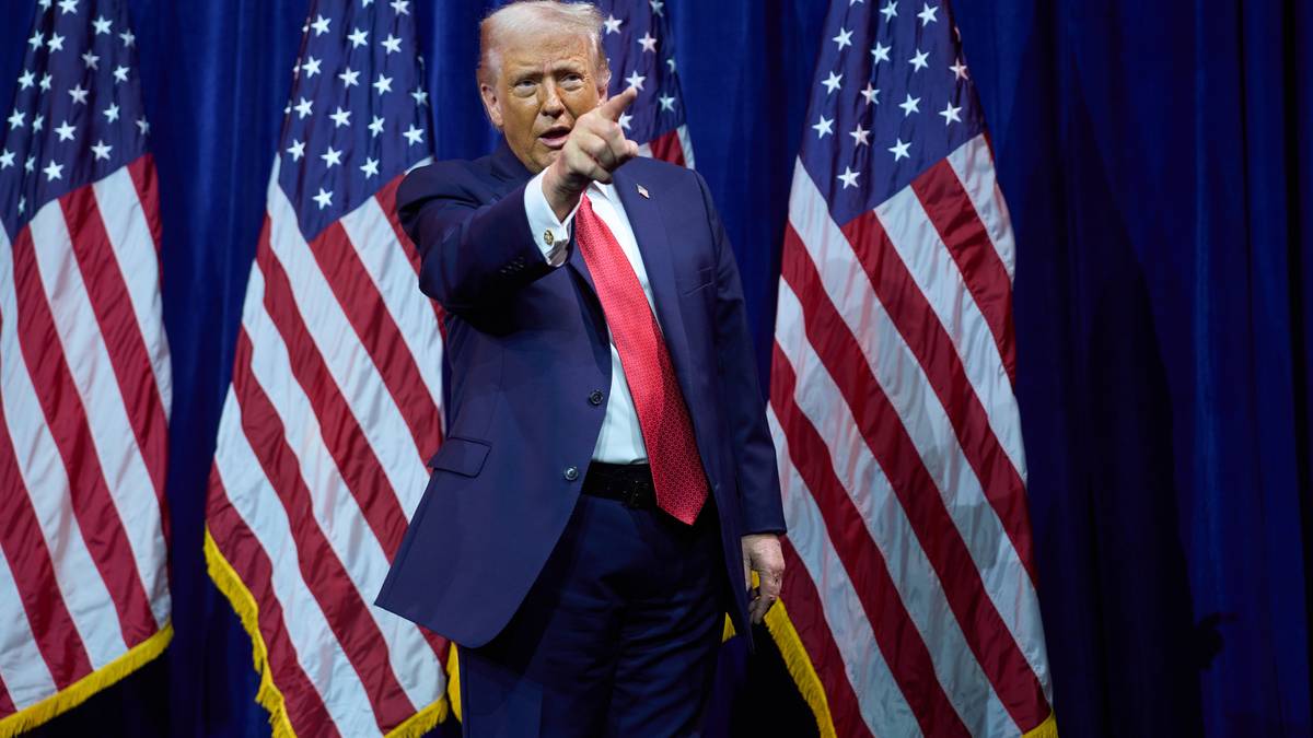 Donald Trump w garniturze i krawacie, stojący przed amerykańskimi flagami i wskazujący palcem.