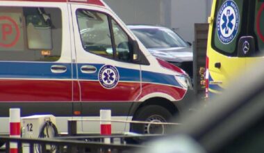 Biało-czerwono-niebieska karetka z symbolem ratunkowym na drzwiach, obok fragment ż&oacute;łtej karetki