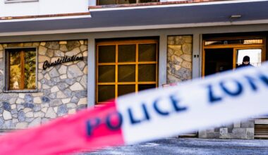 Budynek z kamienną fasadą, z napisem "Constellation" po lewej stronie. Na pierwszym planie widoczna jest taśma policyjna z napisem "POLICE ZONE". W tle, w otwartych drzwiach budynku, stoi osoba ubrana w ciemną kurtkę.