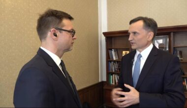 Dw&oacute;ch mężczyzn w garniturach rozmawia w pomieszczeniu z biblioteczką.