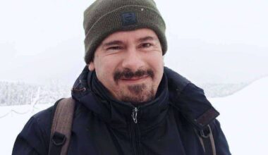 Mężczyzna w zimowej kurtce i czapce z plecakiem na ramionach, uśmiecha się na tle zimowego krajobrazu.