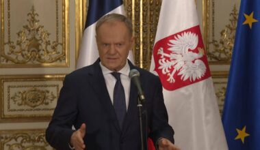 Premier Donald Tusk przemawia przy mikrofonie na tle flagi Polski i Unii Europejskiej.