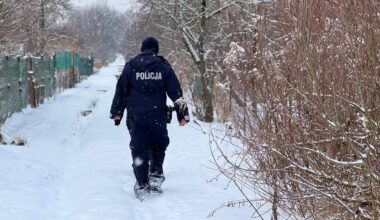 Policjant w mundurze idzie przez zaśnieżoną ścieżkę. Na plecach munduru widoczny napis "POLICJA". Otoczenie jest zimowe, z drzewami pokrytymi śniegiem.