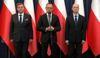 Trzech mężczyzn w garniturach stoi na tle biało-czerwonych flag Polski.