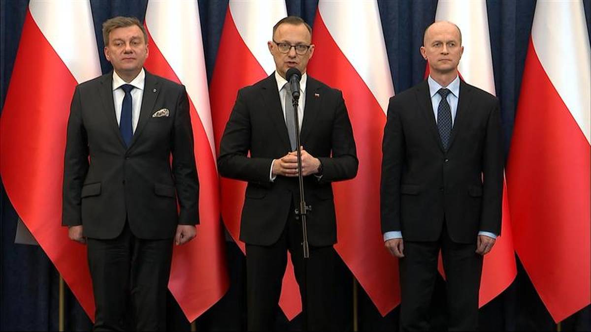 Trzech mężczyzn w garniturach stoi na tle biało-czerwonych flag Polski.