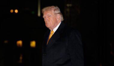 Donald Trump w czarnym płaszczu i ż&oacute;łtym krawacie idzie po chodniku nocą.