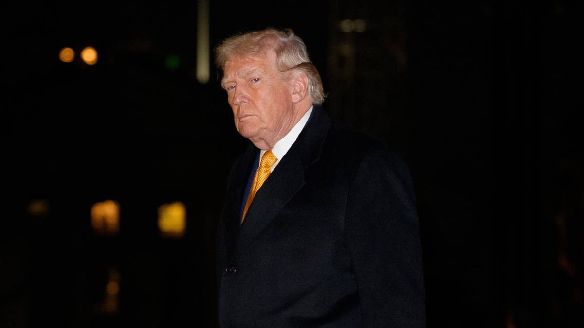 Donald Trump w czarnym płaszczu i ż&oacute;łtym krawacie idzie po chodniku nocą.