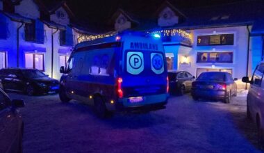 Nocna scena z zaparkowanymi samochodami i ambulansami przed oświetlonym budynkiem.