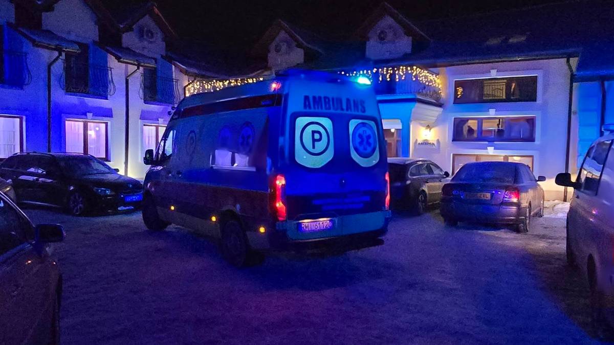 Nocna scena z zaparkowanymi samochodami i ambulansami przed oświetlonym budynkiem.