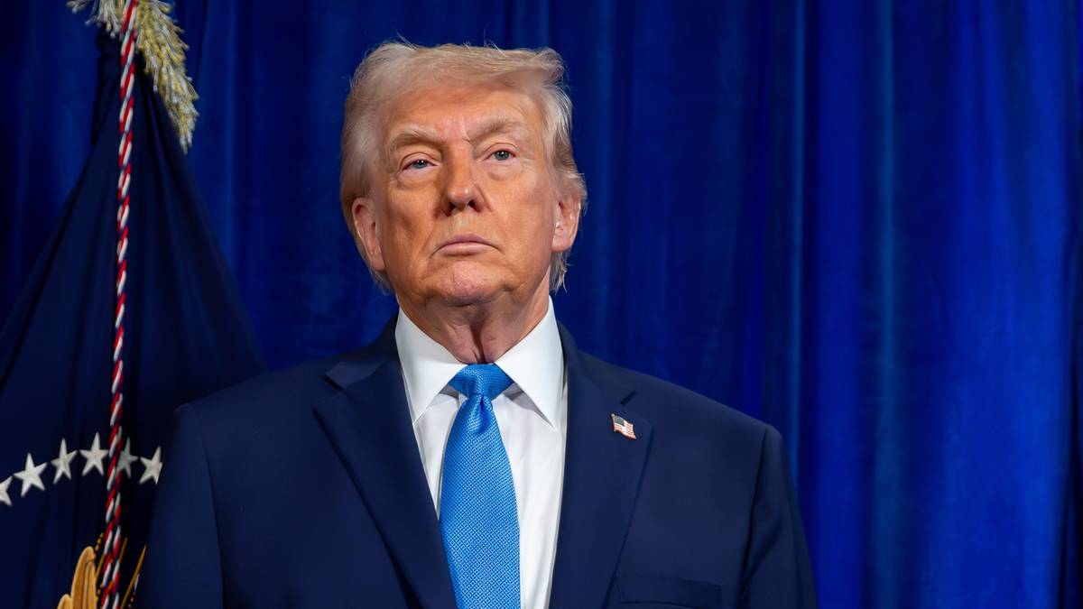 Donald Trump w garniturze i niebieskim krawacie stoi przed niebieską zasłoną.