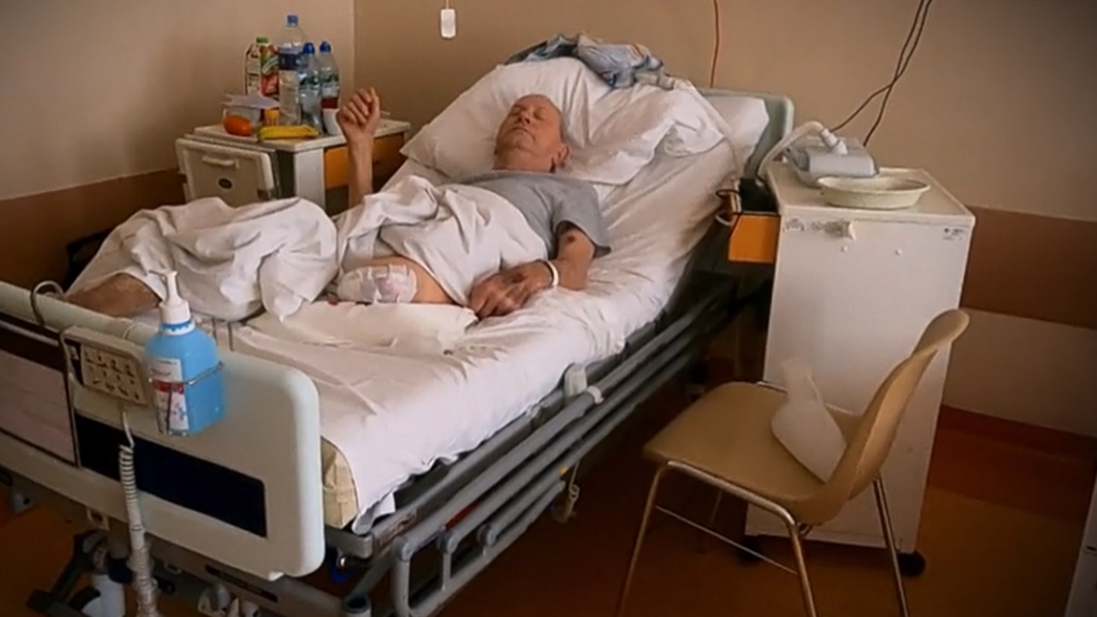 Starszy mężczyzna w szpitalnym ł&oacute;żku, z opatrunkiem na nodze.