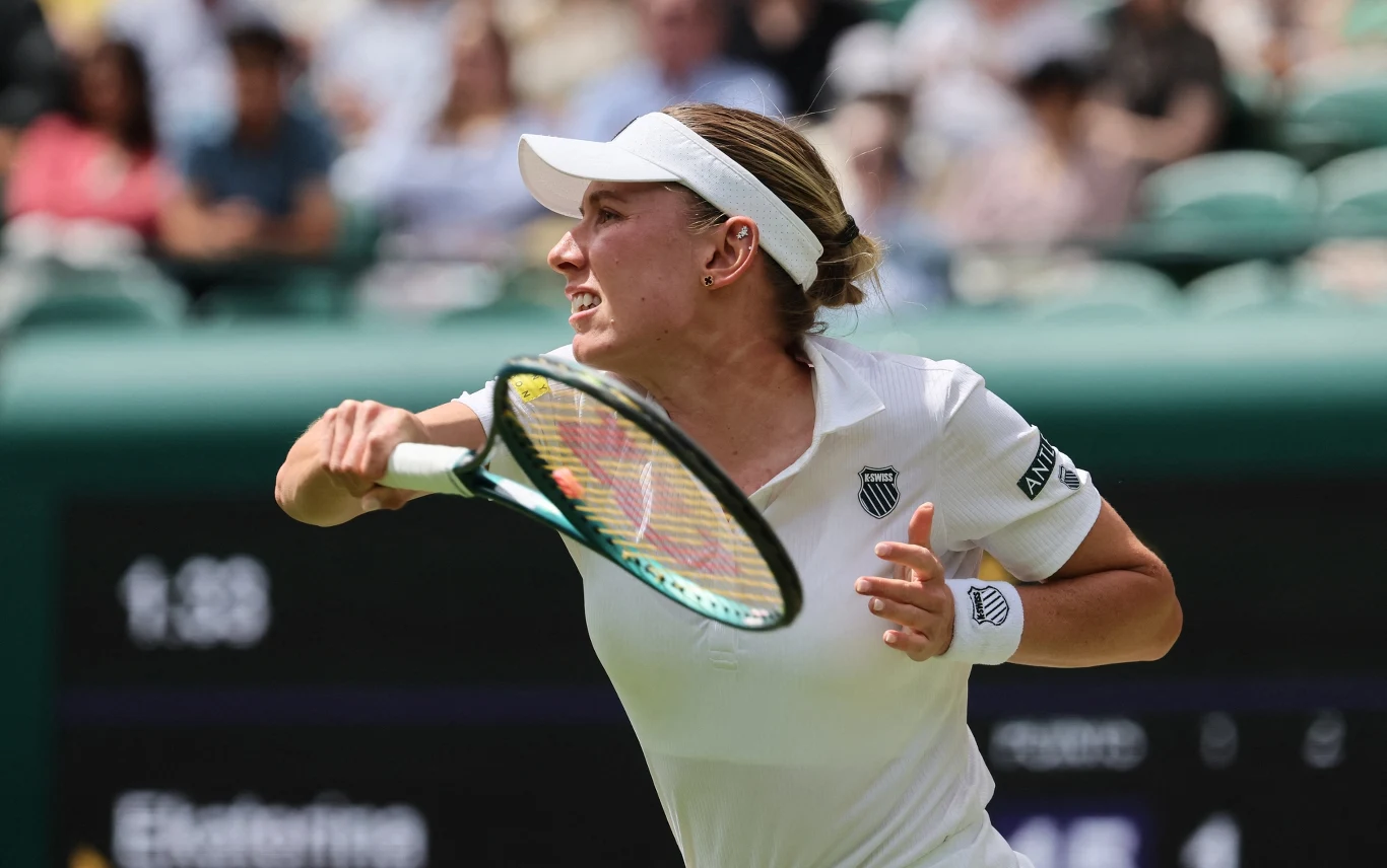 Tenisistka w białym stroju uderza piłkę podczas meczu tenisowego, skupiona i dynamiczna sylwetka z rakietą uniesioną w powietrzu, rozmazane trybuny i widownia w tle.