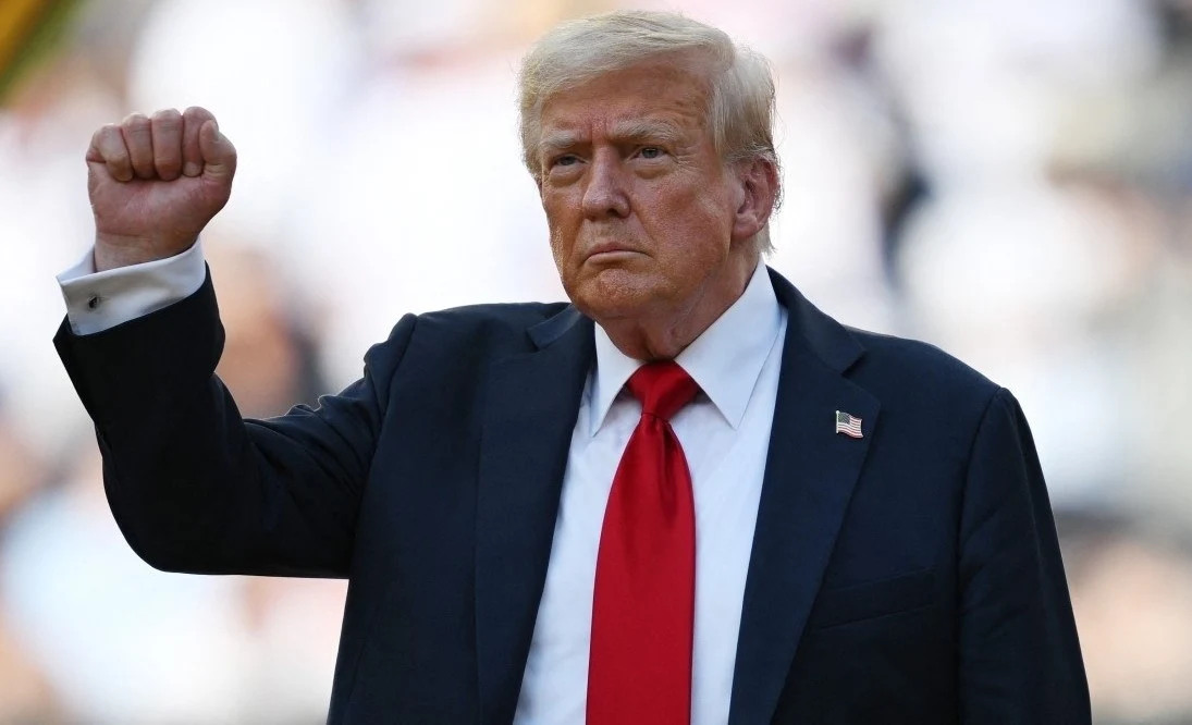 Mężczyzna w garniturze z czerwoną krawatą stoi z zaciśniętą pięścią uniesioną w górę, na jego marynarce widoczna przypinka z flagą USA, tło rozmyte i jasne.