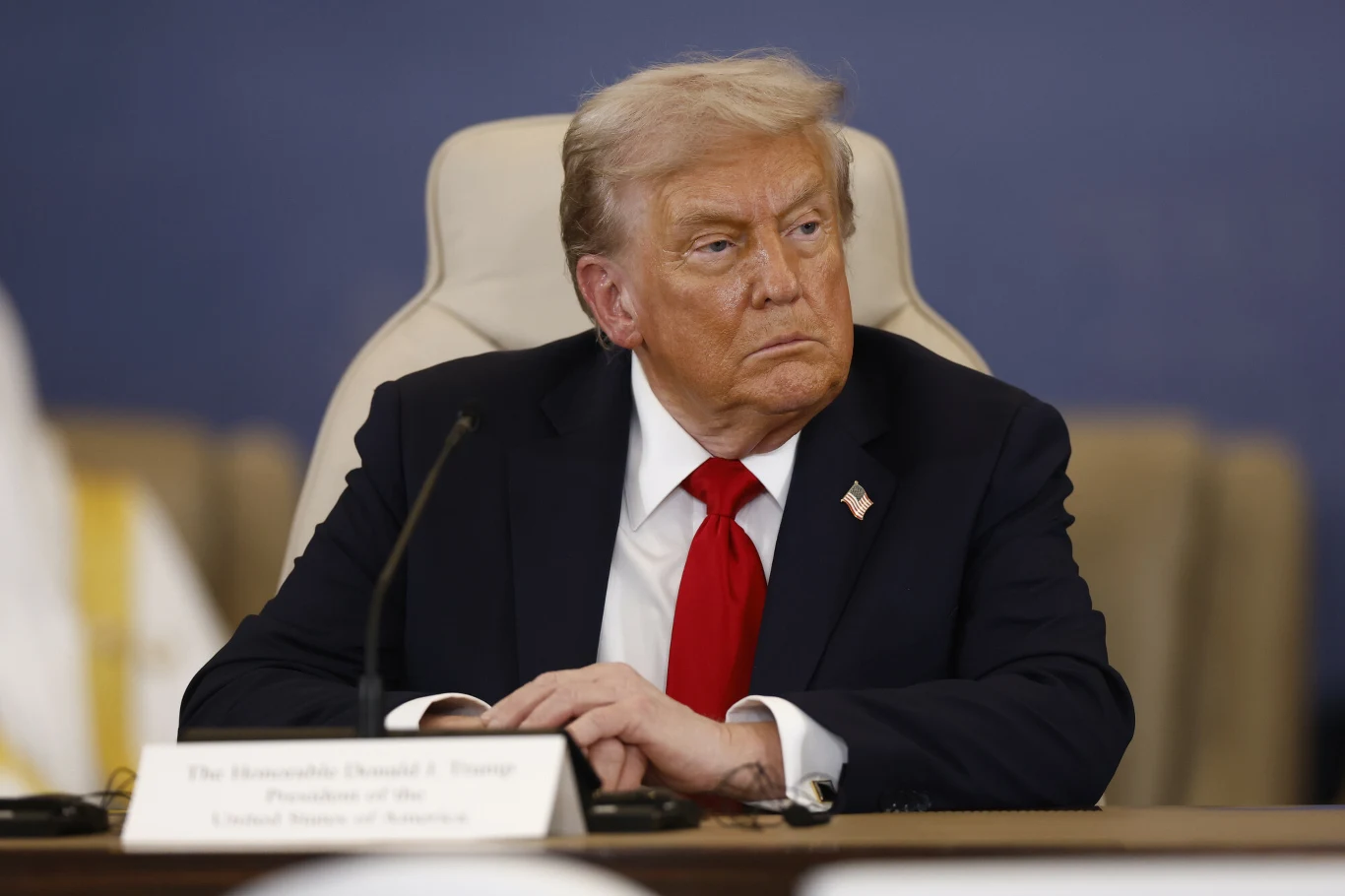 Mężczyzna w średnim wieku w garniturze i czerwonym krawacie siedzi przy stole konferencyjnym, z postawionymi na nim rękami, wyrażając powagę i skupienie. Przed nim leży tabliczka z imieniem i nazwiskiem. W tle neutralne, stonowane kolory.