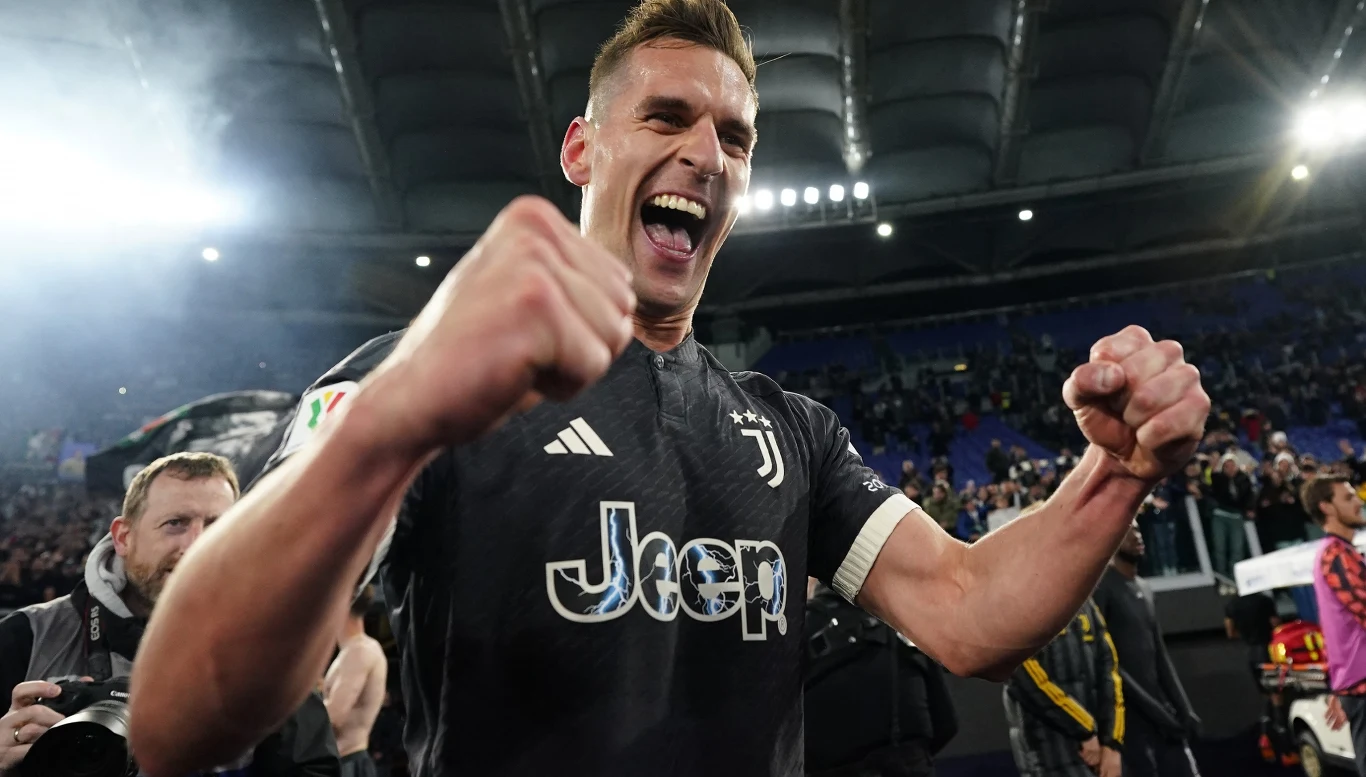 Arkadiusz Milik Piłkarz w koszulce Juventusu z uniesionymi pięściami wyraża ogromną radość na tle stadionu wypełnionego kibicami, wokół znajdują się osoby związane z drużyną i obsługą meczu.