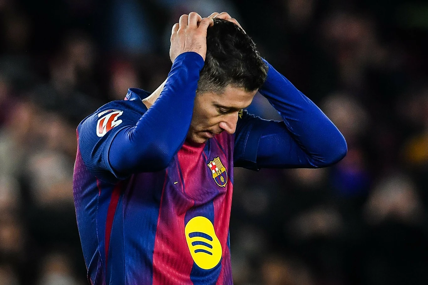 Piłkarz w stroju drużyny FC Barcelona trzyma ręce na głowie, wyrażając rozczarowanie lub frustrację. Jego twarz zwrócona jest w dół, a tło stanowią nieostre sylwetki publiczności.