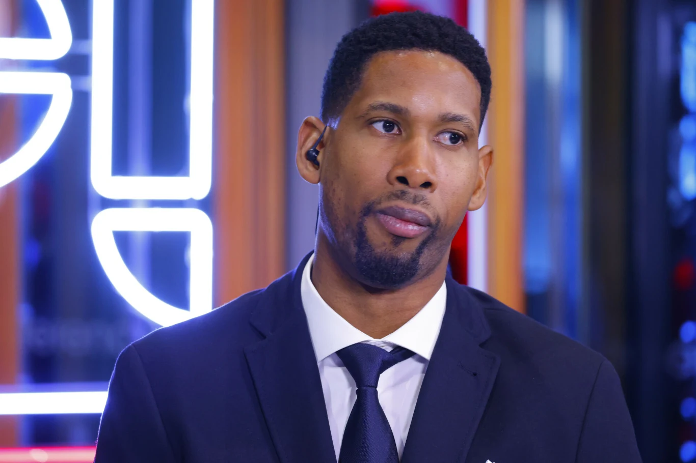 Mężczyzna w eleganckim garniturze i krawacie stoi na tle nowoczesnych, podświetlonych elementów dekoracyjnych. Ma słuchawkę w uchu i neutralny wyraz twarzy.