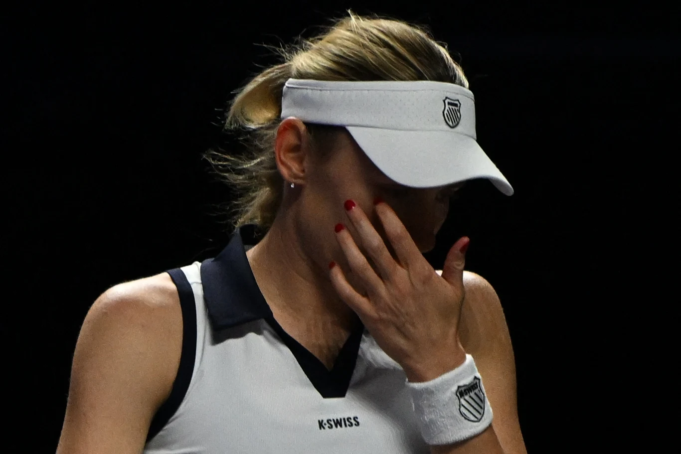 Tenisistka w białym stroju sportowym i daszku zasłania twarz dłonią, wyrażając emocje. Czarne tło skupia uwagę na sylwetce i geście zawodniczki.