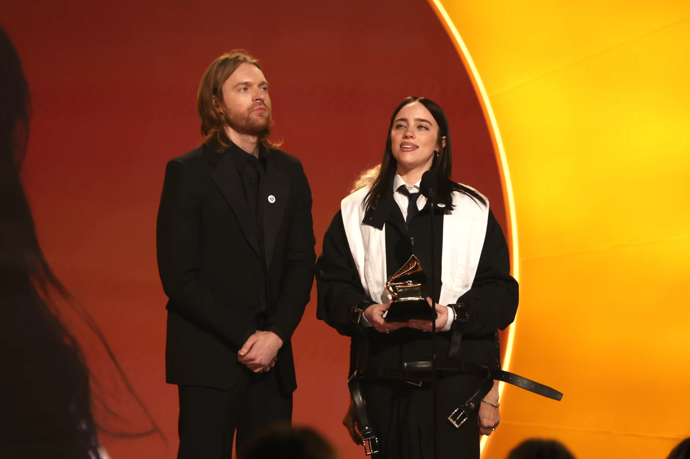 Billie Eilish i jej brat Finneas na scenie Grammy 2026 Dwoje ludzi elegancko ubranych stoi na jasnym tle, jedna z osób trzyma statuetkę przypominającą nagrodę muzyczną Grammy, co sugeruje uroczystą galę wręczenia nagród.