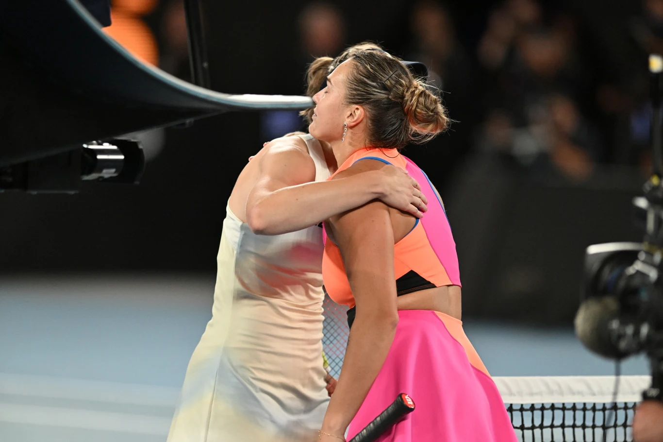 Dwie tenisistki przy siatce obejmują się po zakończeniu meczu na korcie, jedna z nich trzyma rakietę tenisową, obie ubrane w sportowe stroje, w tle widać rozmytą publiczność.