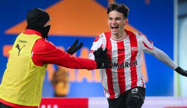 Piłkarz w biało-czerwonej pasiasto-czerwonej koszulce z radością biegnie po murawie, wykonując gest triumfu, w tle rozmazane postaci i elementy stadionu.