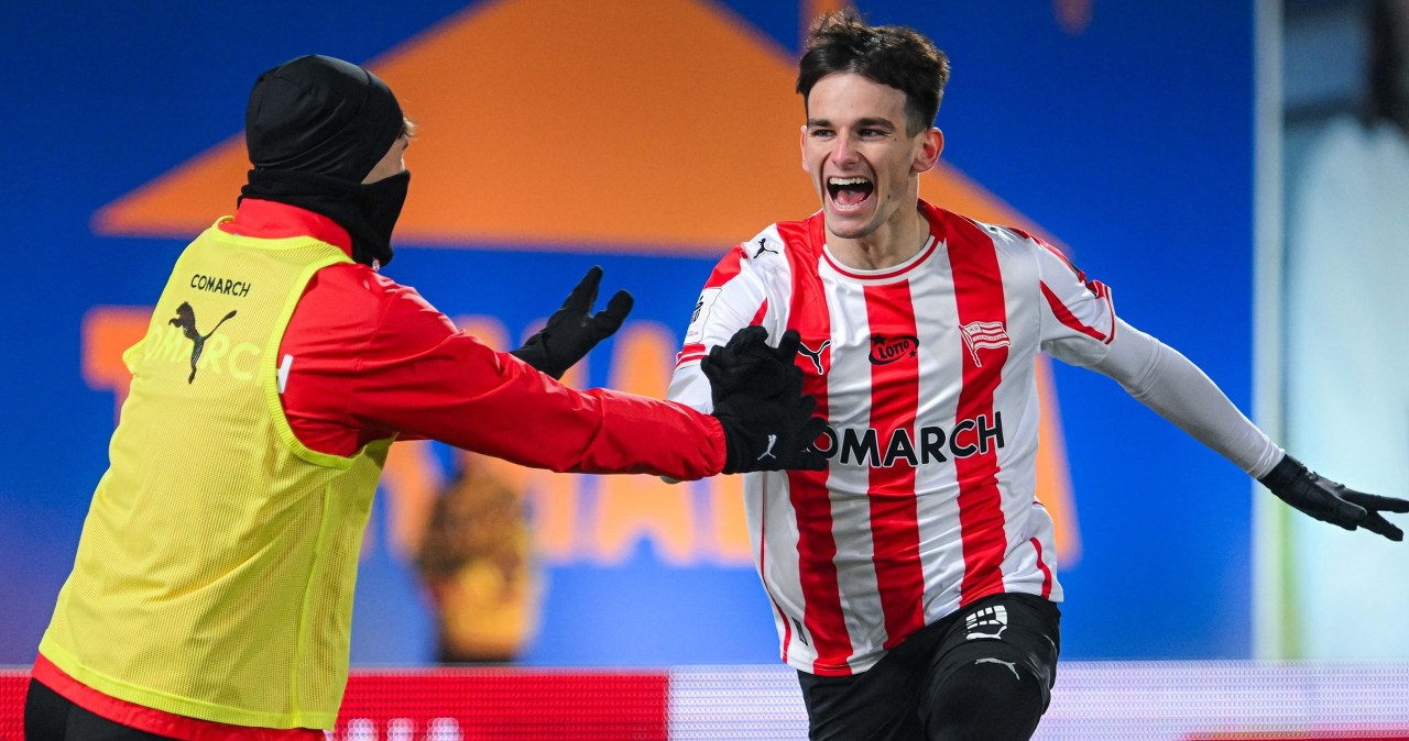 Piłkarz w biało-czerwonej pasiasto-czerwonej koszulce z radością biegnie po murawie, wykonując gest triumfu, w tle rozmazane postaci i elementy stadionu.