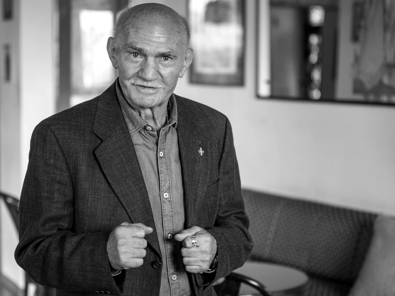 Starszy mężczyzna z siwymi włosami i wąsami, ubrany w elegancką marynarkę i koszulę, stoi w jasnym pomieszczeniu z wygodnymi meblami w tle. Trzyma dłonie uniesione przed sobą w geście jakby chciał coś pokazać lub podkreślić wypowiedź. Na twarzy wyraz ż...