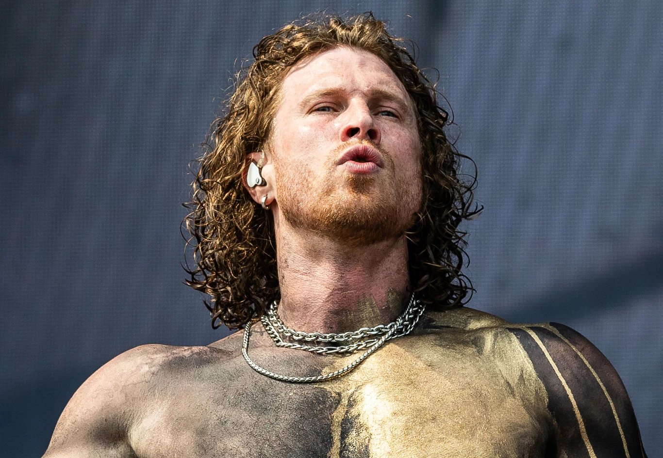 Mężczyzna z długimi, kręconymi włosami i zarostem, z łańcuchem na szyi, ma pokryte farbą ciało od ramion do klatki piersiowej, stoi na tle rozmazanego, szarego tła.