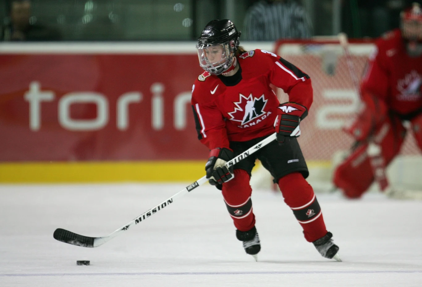 Zawodniczka w czerwonym stroju drużyny Kanady prowadzi krążek hokejowy na lodzie podczas meczu, w tle widoczne są elementy lodowiska oraz bramkarz.