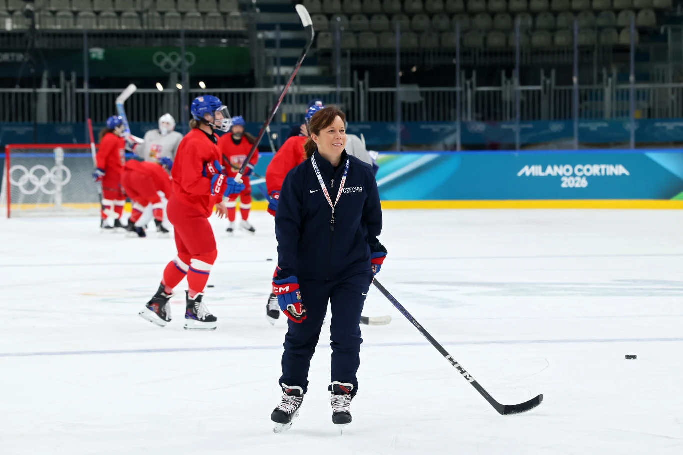 Trenerka hokejowa na lodzie z kijem, w tle kilku zawodników w czerwonych strojach ćwiczy na lodowisku podczas przygotowań do igrzysk olimpijskich w Mediolanie Cortina 2026.