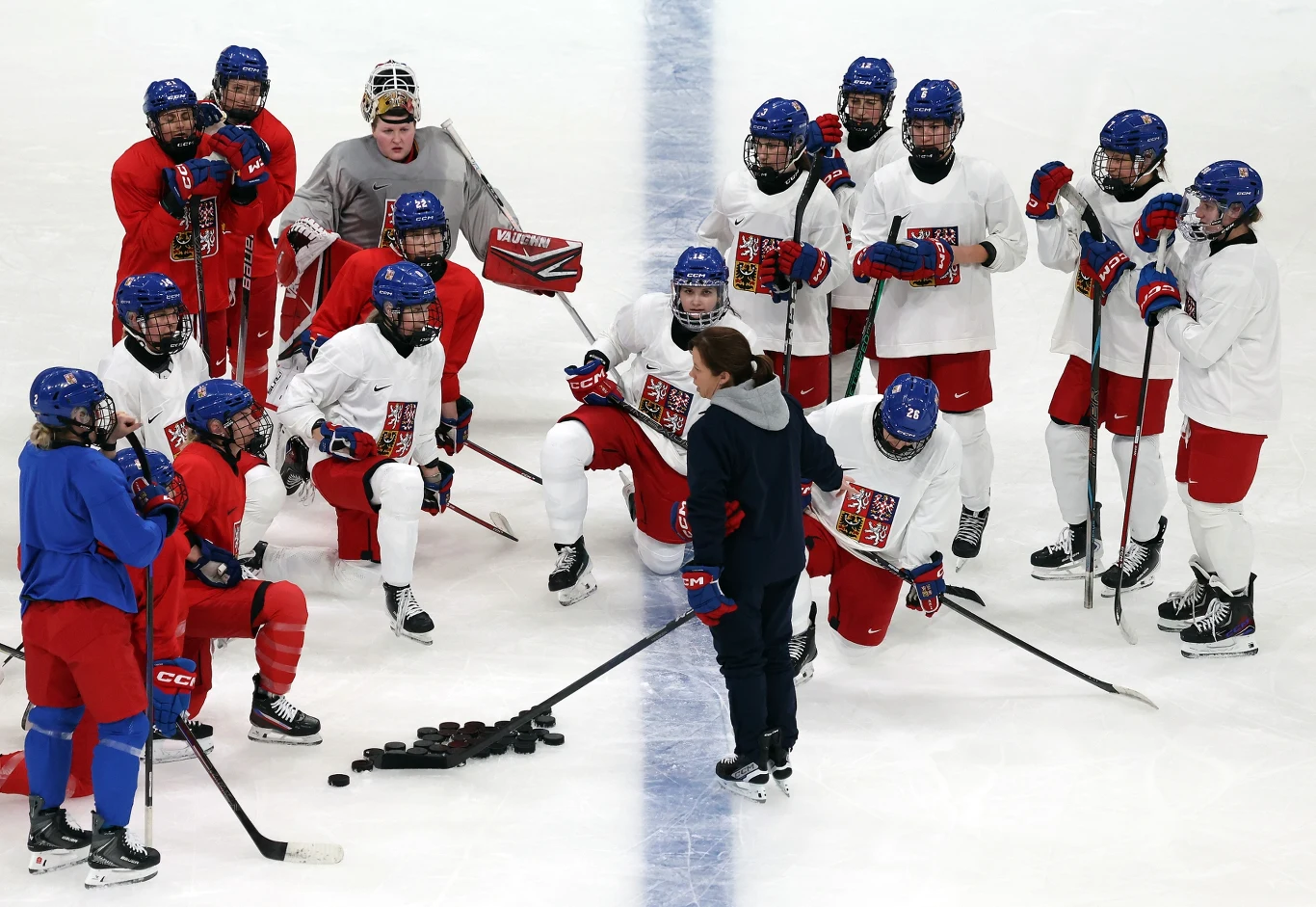 Grupa hokeistów na lodzie w strojach treningowych słucha trenera, który tłumaczy ćwiczenie, przed nimi na lodzie leży kilka kijów hokejowych oraz stos krążków.
