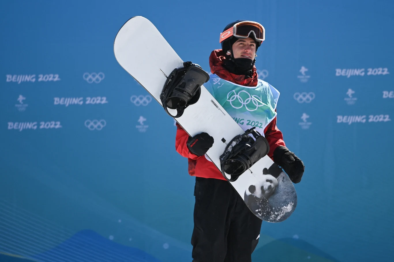 Snowboardzista w czerwonej kurtce trzymający deskę snowboardową, w tle niebieskie tło z logotypami Zimowych Igrzysk Olimpijskich oraz napisem Beijing 2022.
