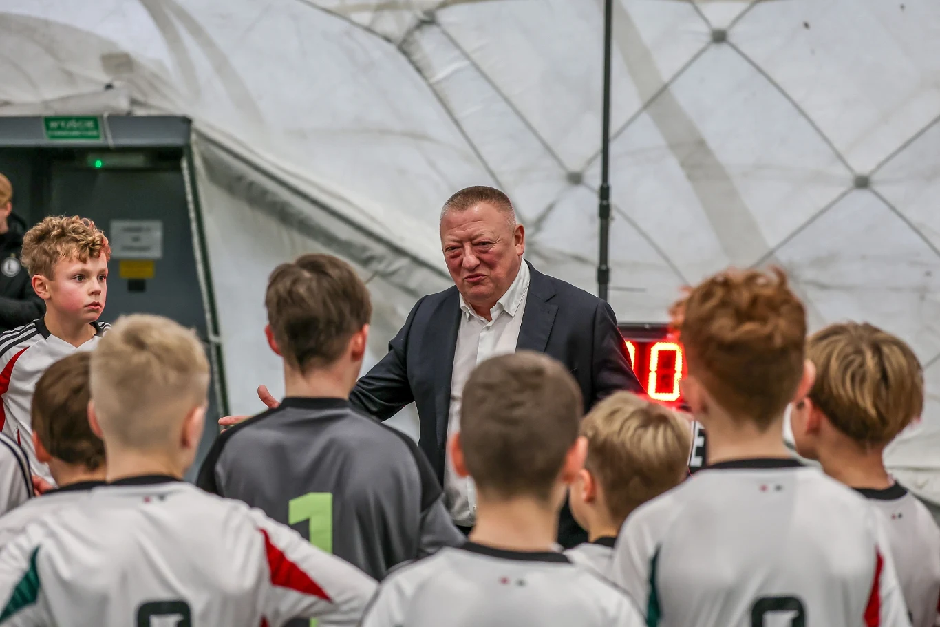 Mężczyzna w garniturze przemawia do grupy młodych chłopców w strojach sportowych stojących w półkolu na hali sportowej z widocznym elektronicznym zegarem w tle.