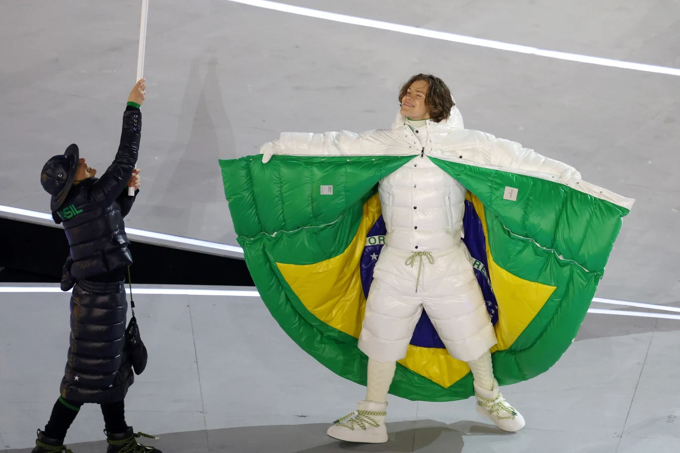 Lucas Braathen podczas ceremonii otwarcia IO 2026 Osoba ubrana w biały strój otwiera kurtkę wyłożoną w środku brazylijską flagą, obok stoi druga osoba z flagą na kiju. Sytuacja rozgrywa się na stadionie, podkreślając elementy sportowe i narodowe.