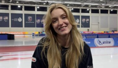 Portret uśmiechniętej młodej kobiety z długimi blond włosami, siedzącej w obiekcie sportowym z lodowiskiem w tle.