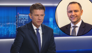 Mężczyzna w garniturze w studiu telewizyjnym obok portretu innego mężczyzny w garniturze.