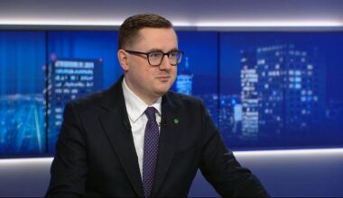 Mężczyzna w garniturze i okularach siedzi przed ekranami z nocnym widokiem miasta.