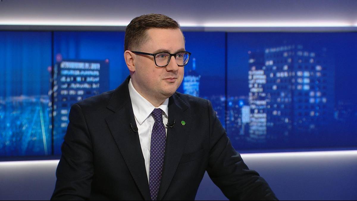 Mężczyzna w garniturze i okularach siedzi przed ekranami z nocnym widokiem miasta.