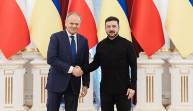 Premier Polski Donald Tusk i Prezydent Ukrainy Wołodymyr Zełenski ściskają sobie dłonie na tle flag Polski i Ukrainy.