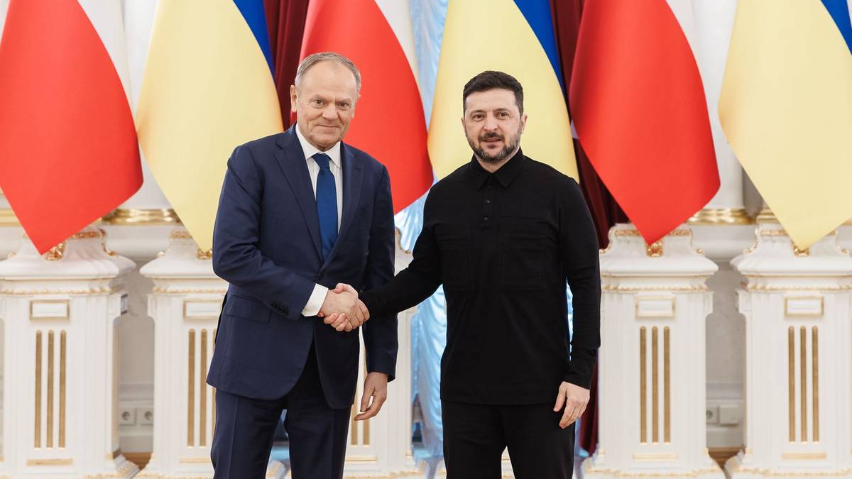Premier Polski Donald Tusk i Prezydent Ukrainy Wołodymyr Zełenski ściskają sobie dłonie na tle flag Polski i Ukrainy.