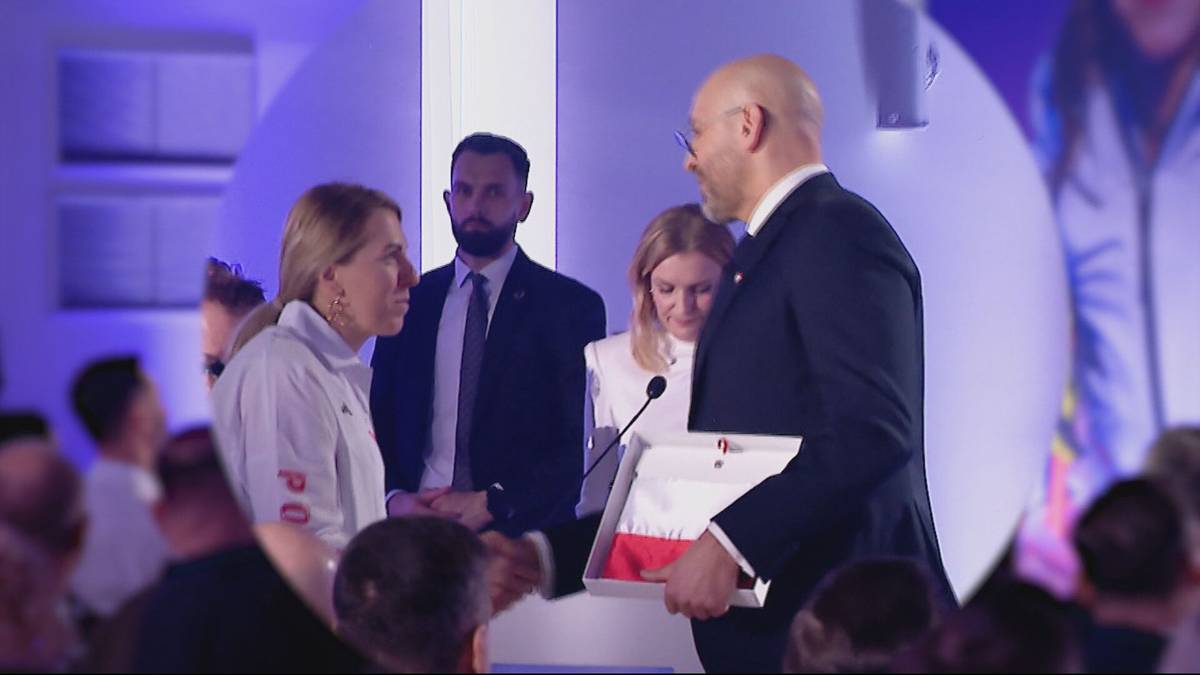 Mężczyzna w garniturze wręcza kobiecie dokument w ramce podczas oficjalnej ceremonii.