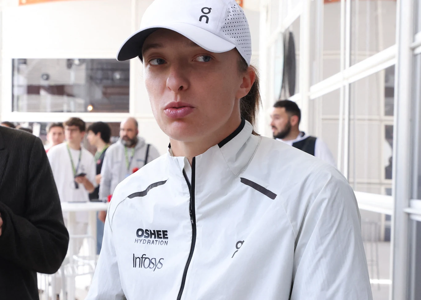 Iga Świątek Sportowiec w białej kurtce i czapce z daszkiem, ubrany w odzież z widocznymi logotypami sponsorów, stoi w jasnym, nowoczesnym wnętrzu pośród kilku osób w tle.